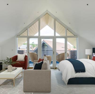 A spacious bedroom with a vaulted ceiling features a bed, a TV, a red couch, a beige loveseat, and a small balcony with outdoor chairs, all under large triangular windows.