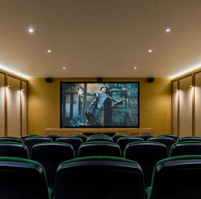 A small home theater with green seating faces a large screen displaying an action movie scene. Soft lighting and beige walls complete the cozy setup.