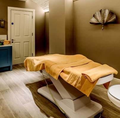 A massage room with a treatment table, covered with a yellow towel, a sink with toiletries, and decorative wall fans.