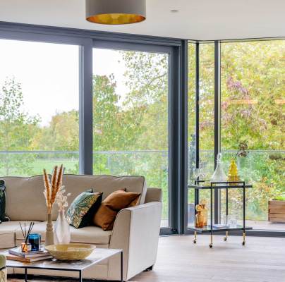 Bright living room with large windows, a beige sofa with colorful pillows, a glass and metal side table, and an armchair. Greenery and trees visible outside.