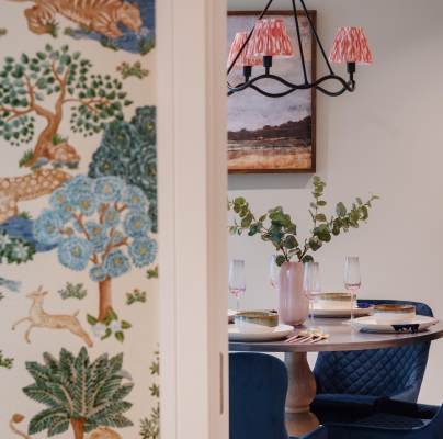 A dining area with a round table set for a meal, four blue chairs, and a vase with leaves. A wall with a colorful nature-themed wallpaper is partially visible.