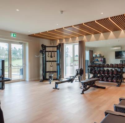 A modern gym with exercise machines, weights, and workout benches. Large windows and a mirrored wall enhance the spacious interior.