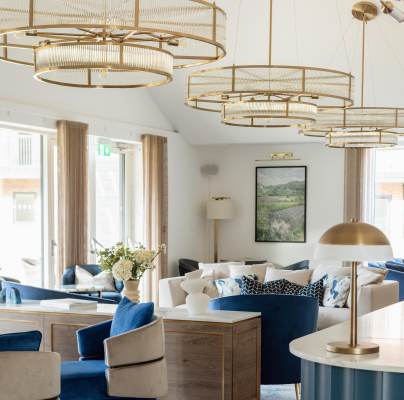 Spacious modern lounge with large windows, blue and white seating, gold lighting fixtures, and a central table with flowers.