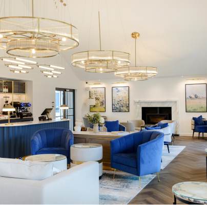 Elegant hotel lounge with blue and white sofas, marble tables, and modern chandeliers. A bar with coffee machines is in the corner. Large artworks and a fireplace adorn the walls.