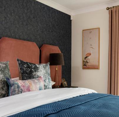 Bedroom with a bed featuring a blue quilt, patterned pillows, a brown headboard, a black lamp on a side table, dark textured wallpaper, and a framed bird print. Curtains cover a window.