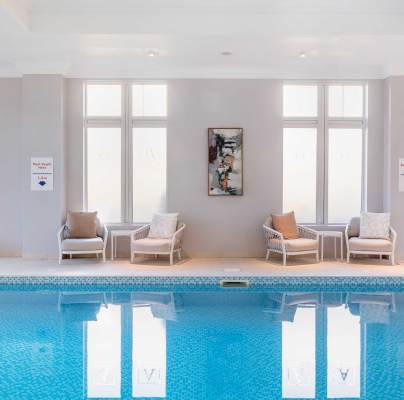 Indoor swimming pool with clear water, surrounded by white walls and large windows. Two chairs with cushions and a wall art piece are visible on the right side.