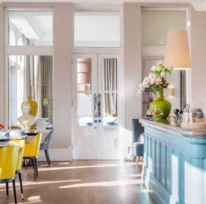 Bright cafe interior with yellow chairs, wooden tables, and a blue counter. Large windows allow natural light, while decorative vases and flowers add charm.