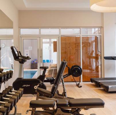 A gym with various equipment including dumbbells, exercise bikes, and treadmills. A mirrored wall and a view of a pool area through glass doors in the background.