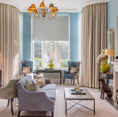 Elegant living room with blue walls, large windows, and beige curtains. Features a chandelier, fireplace, sofas, armchairs, and decorative sculptures. A framed painting hangs on the wall.