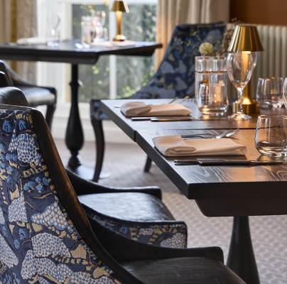 Elegant dining setup with tables and chairs. Tables have black surfaces, set with napkins, cutlery, glasses, and small lamps. Chairs have a blue patterned design. Soft natural light from a window.