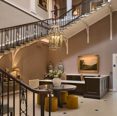 Elegant hotel lobby with a grand staircase, chandelier, art on walls, round table with flowers, and seating area. Neutral tones dominate the decor. A doorway leads to a room labeled "The Morning Room.