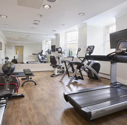 A gym with treadmills, stationary bikes, a rowing machine, free weights, and benches on a wooden floor. Mirrors and a wall-mounted TV are also visible.