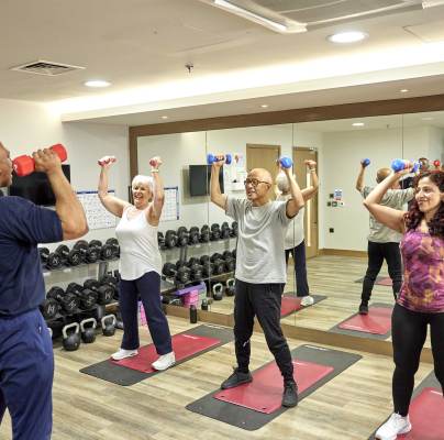 A fitness instructor guides three people lifting dumbbells in a gym mirrored room.