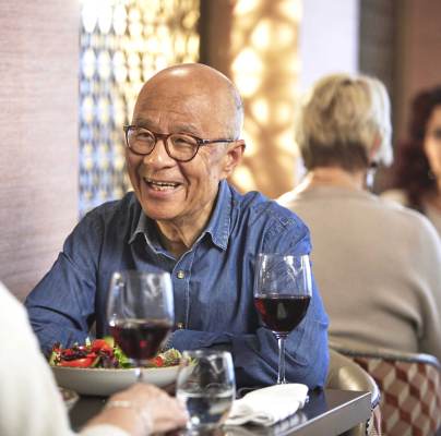 Older adults dining at a restaurant, engaging in conversation. Two glasses of red wine and a salad are on the table.