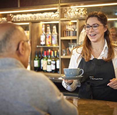 Barista in an apron serves a cup to a seated customer at a counter. Shelves with bottles and a coffee machine are in the background.