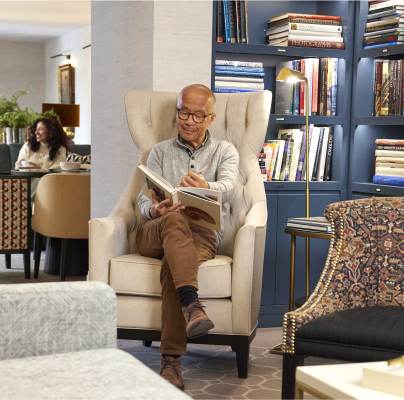 A man sits in an armchair reading a book in a library-like room. Shelves filled with books are visible in the background, and two people converse at a nearby table.