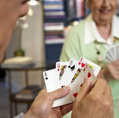 Two people playing cards at a table. One person holds a hand with an ace of clubs, queen of hearts, five of spades, king of hearts, and four of hearts. The other person smiles, holding their cards.