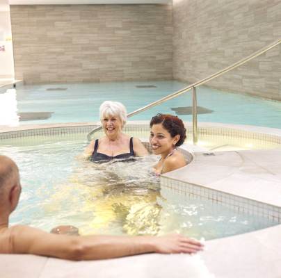 Three people relaxing in a hot tub at an indoor pool facility. Two women are seated in the water, facing a man who's sitting on the tub's edge.