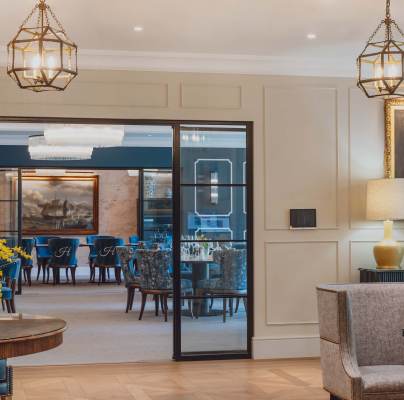 Elegant interior with modern lighting, a round table with flowers, and a large portrait on the wall. Glass doors open to a dining area with blue chairs and a painting.