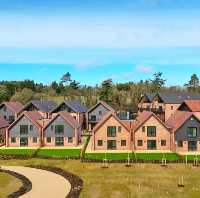 A row of modern brick houses with red roofs and manicured lawns, set against a backdrop of trees and blue sky.