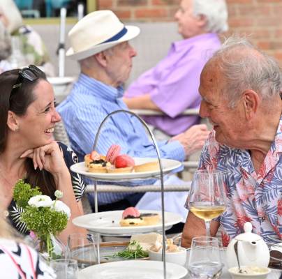Two people sit at a table outdoors, smiling and talking, with plates of desserts and glasses of wine in front of them. Other diners are visible in the background.