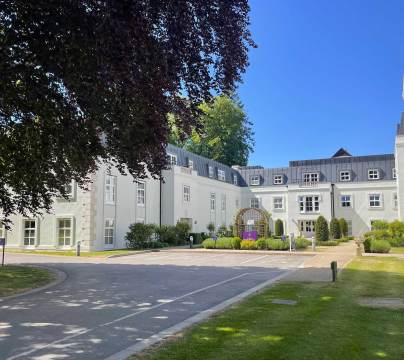 White multi-story building with black roof, surrounded by green lawn, trees, and a driveway under a clear blue sky.