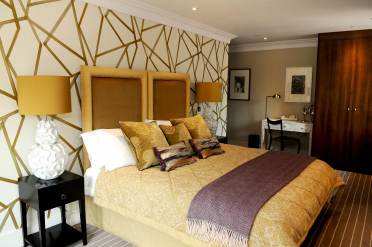 A bedroom with a double bed, white and yellow table lamps on the sides and white walls with golden lines