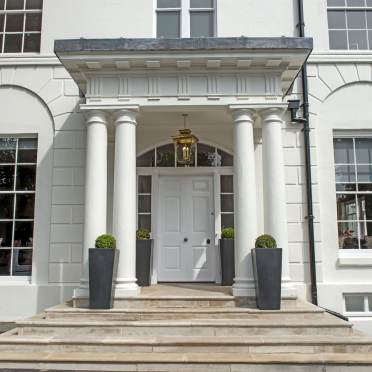 White front entrance of a building with four columns, a central door, two windows, potted plants on each side, and a hanging lantern above the steps.