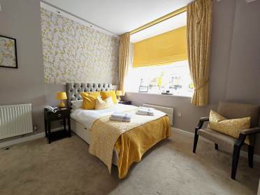 A neatly arranged hotel room with a double bed, yellow and gray decor, a window with yellow curtains, and a chair beside the bed. Towels are placed on the bed.