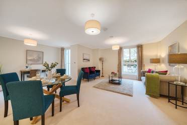 Spacious living and dining area with blue chairs, two couches, a coffee table, and a dining table set. Large windows with curtains let in natural light. Neutral tones and modern decor.