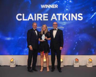 Three people in formal attire stand on a stage. A woman in the middle holds a trophy. A screen behind them displays "Winner Claire Atkins.