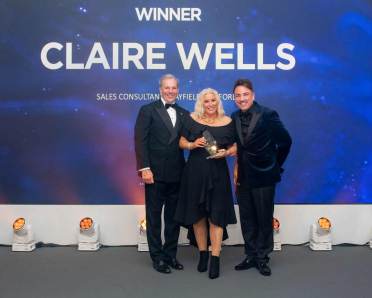 Three people stand on stage, one woman holds an award. The backdrop reads "Winner Claire Wells Sales Consultant." They are dressed in formal attire.