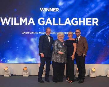 Four people stand on a stage celebrating a winner, with a large background screen displaying the name "Wilma Gallagher.
