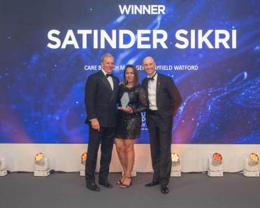 Three people are standing together on a stage. The person in the center is holding an award, with a backdrop displaying "Winner Satinder Sikri.