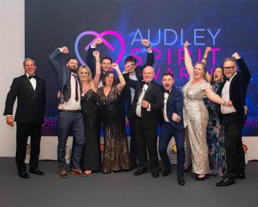 A group of people dressed in formal attire celebrate together at an event, standing in front of a backdrop that reads "Audley Spirit Awards.