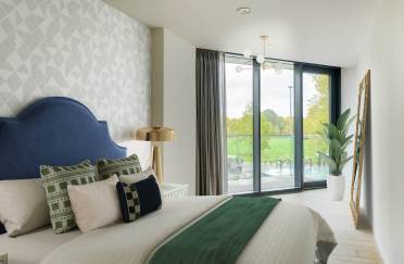 A modern bedroom with a blue headboard, patterned pillows, a brass lamp, and a green throw on the bed. Large windows show a view of trees and a sliding glass door leads to a balcony.