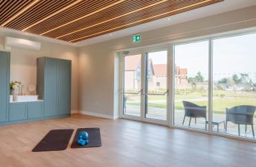 Spacious room with wooden flooring, two exercise mats, a blue kettlebell, and a kitchenette. Large glass doors open to a garden view.