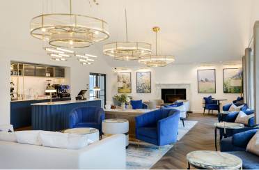 Elegant hotel lounge with blue and white sofas, marble tables, and modern chandeliers. A bar with coffee machines is in the corner. Large artworks and a fireplace adorn the walls.