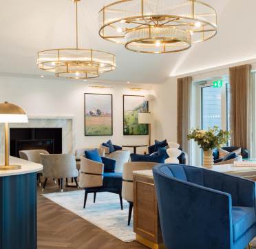 A modern living room with blue and beige furniture, large gold chandeliers, and nature-themed paintings. A fireplace is in the background, and large windows let in natural light.