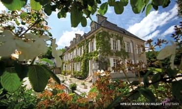 Stone manor covered in ivy with white shutters, surrounded by foliage, set against a sunny sky with scattered clouds. Text at the bottom reads "HPB Le Manoir du Hilguy - Brittany.