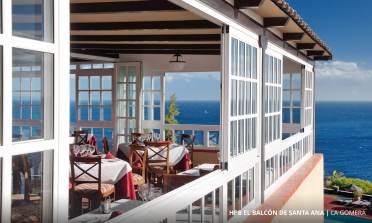 A restaurant with large windows overlooks a scenic ocean view. Tables are set with red tablecloths and place settings.