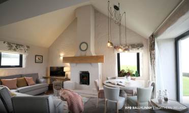 Cozy living room with a fireplace, modern chandelier, sofa, TV, round dining table, and large windows overlooking greenery. White walls and wooden accents add warmth to the space.