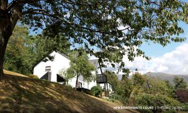 White house on a grassy slope, partially shaded by a large tree. Mountains and blue sky with clouds in the background.