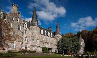 Large stone building with round towers and pointed roofs set against a backdrop of trees and blue sky, labeled "HPB Tigh Mor | The Scottish Highlands.