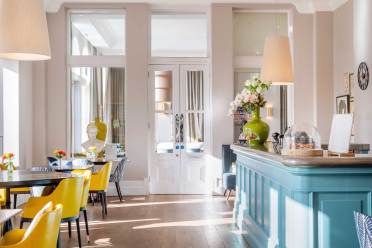 Bright cafe interior with yellow chairs, wooden tables, and a blue counter. Large windows allow natural light, while decorative vases and flowers add charm.