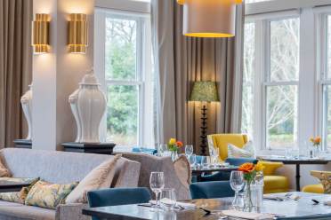 Owners Lounge A modern dining room with chairs, tables set with glassware, and bright natural light from large windows. Vases and lamps decorate the space.