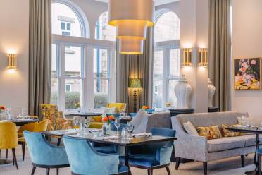 Elegant restaurant interior with arched windows, blue and yellow seating, and tables set with glassware. A contemporary chandelier hangs above, and a framed artwork is on the wall.