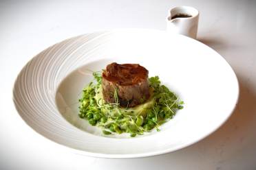 Slow Braised Lamb Henry A gourmet dish featuring a cylinder of cooked meat on a bed of peas and greens, with a sprig garnish, served on a white plate. A small sauce pitcher is in the background.