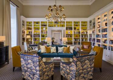 Elegant library room with a chandelier, large yellow bookshelves, a green sofa, patterned armchairs, and a central coffee table, creating a cozy reading area.