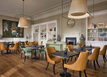 A modern cafe with brown and green chairs, wooden tables, large pendant lights, and wall art. Shelves display decorative vases.